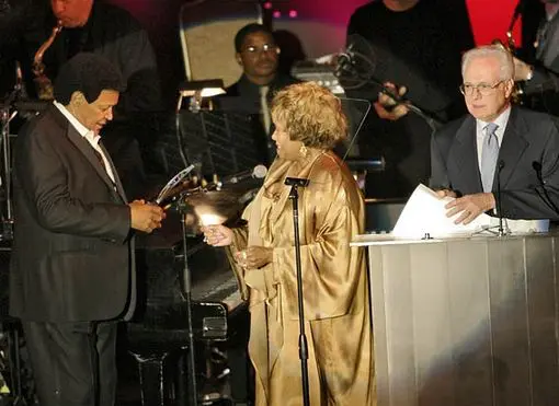 A woman in gold jacket holding microphone and standing next to other people.